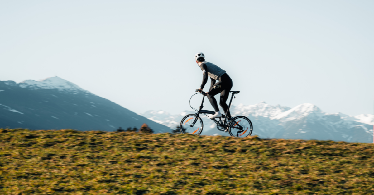 A cyclist rides an e-bike across a grassy hillside with snow-capped mountains in the background.