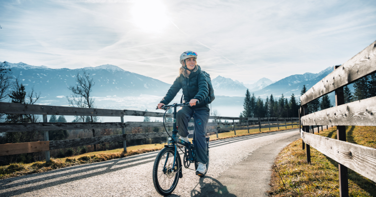 A young person rides a bicycle on a paved path in the countryside, with mountains in the background.