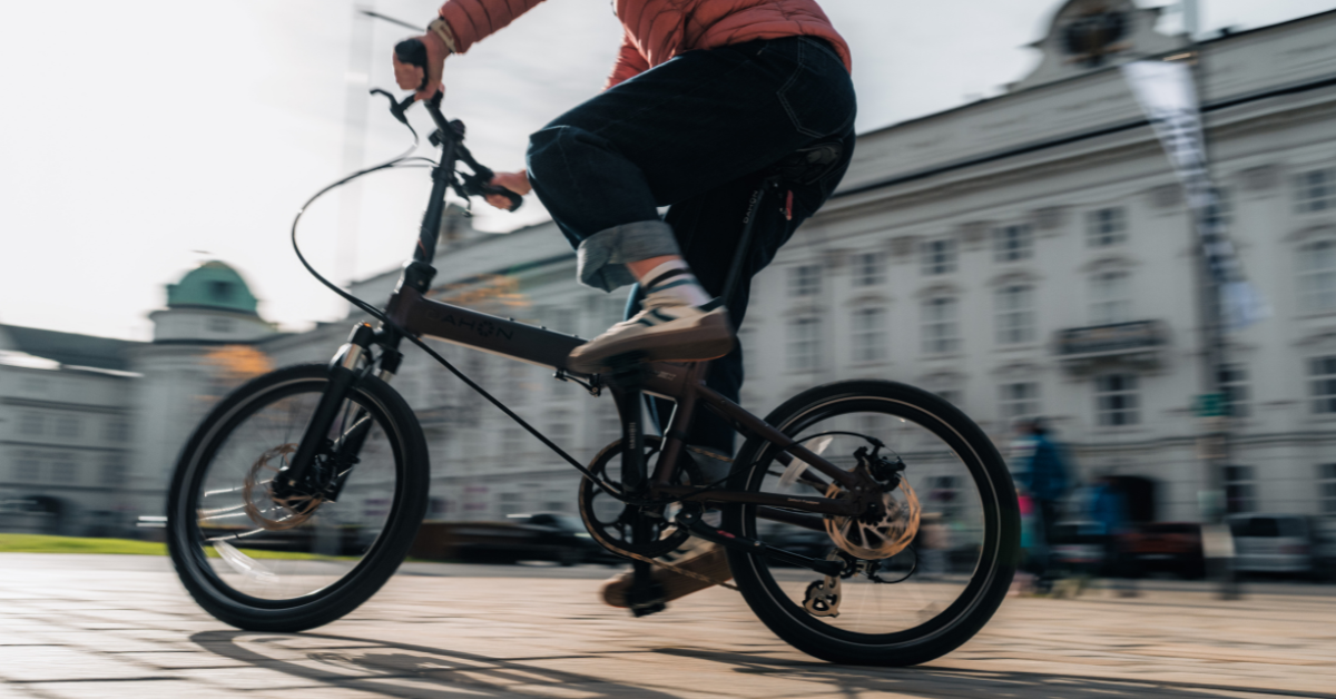 A person rides a black foldable e-bike on a city street, with historic buildings blurred in the background.