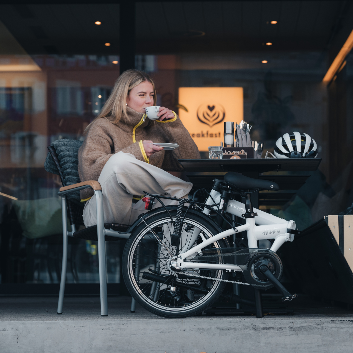 A woman drinking coffee at an outdoor cafe with a folded bike.
