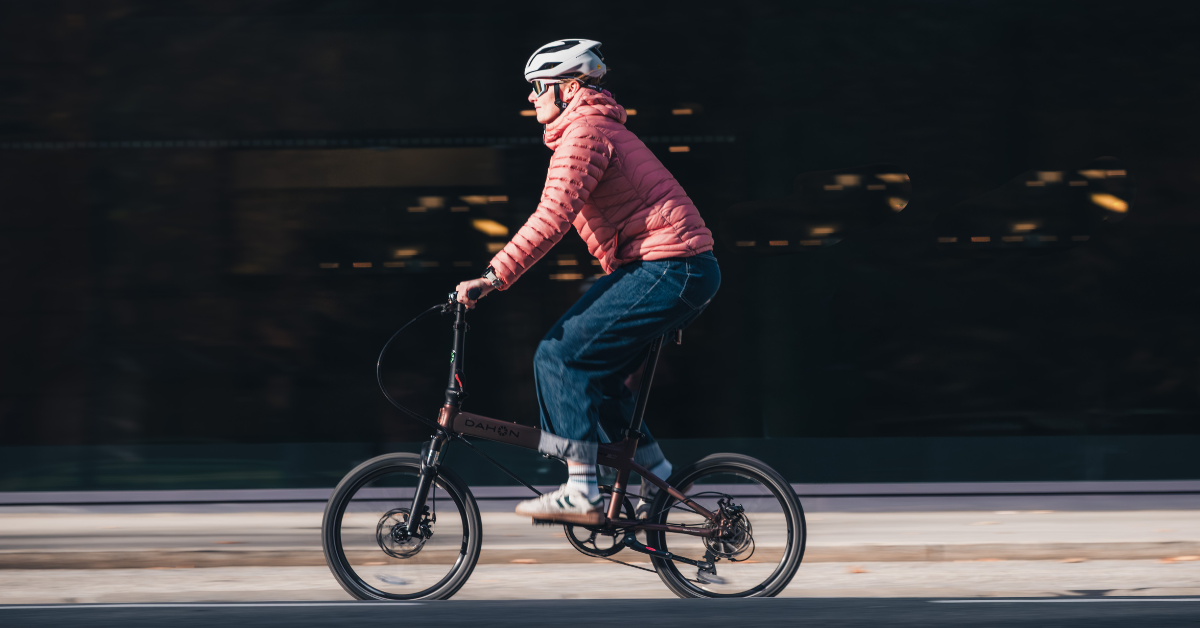 A person in a pink jacket and helmet rides a folding bike along a city street.