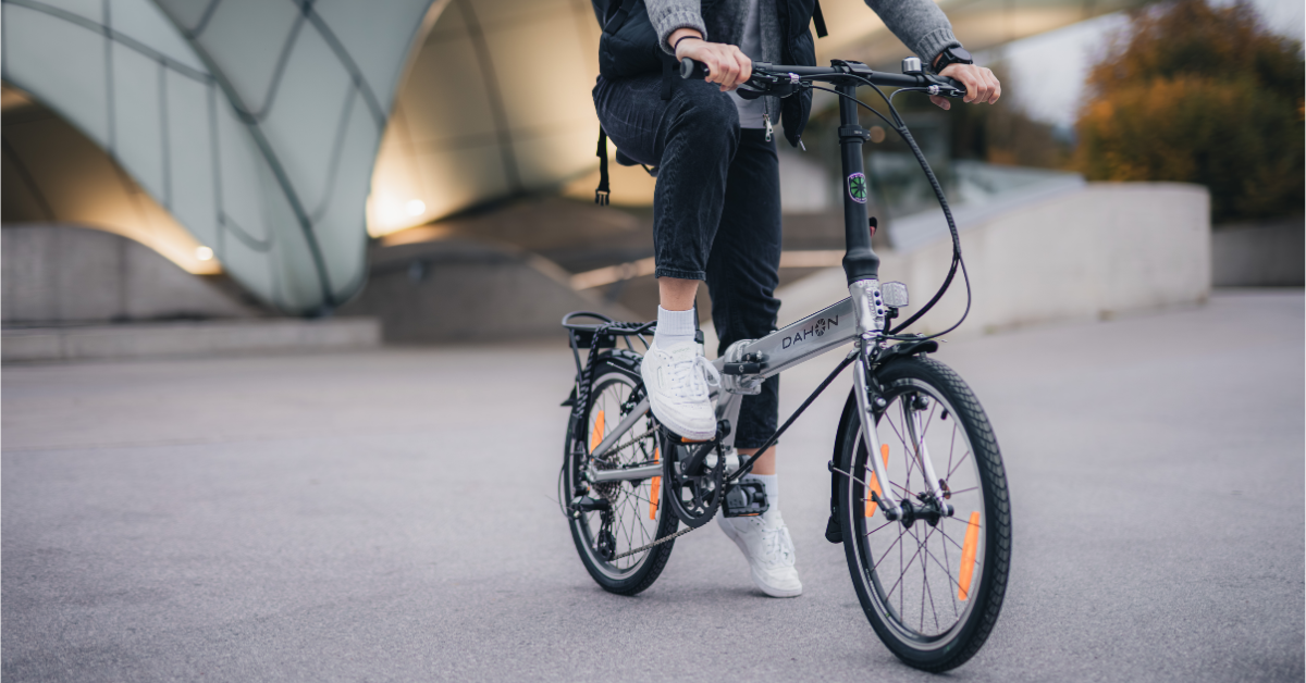A person in casual clothes rides a silver folding bike, with modern architecture in the background.