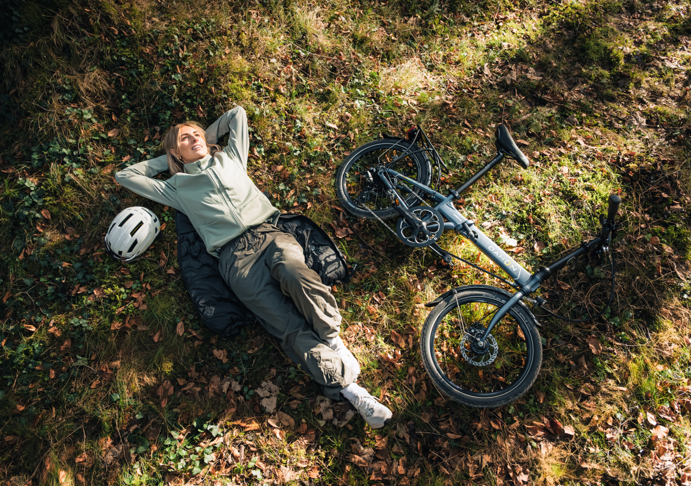 A person resting on forest ground next to a folding bike and helmet.