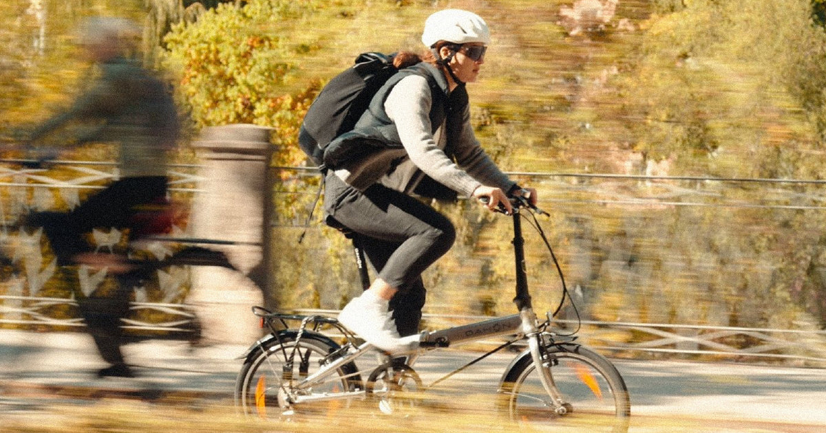 A cyclist wearing a white helmet and black backpack rides a folding bike with motion blur, suggesting speed, against an autumnal background.