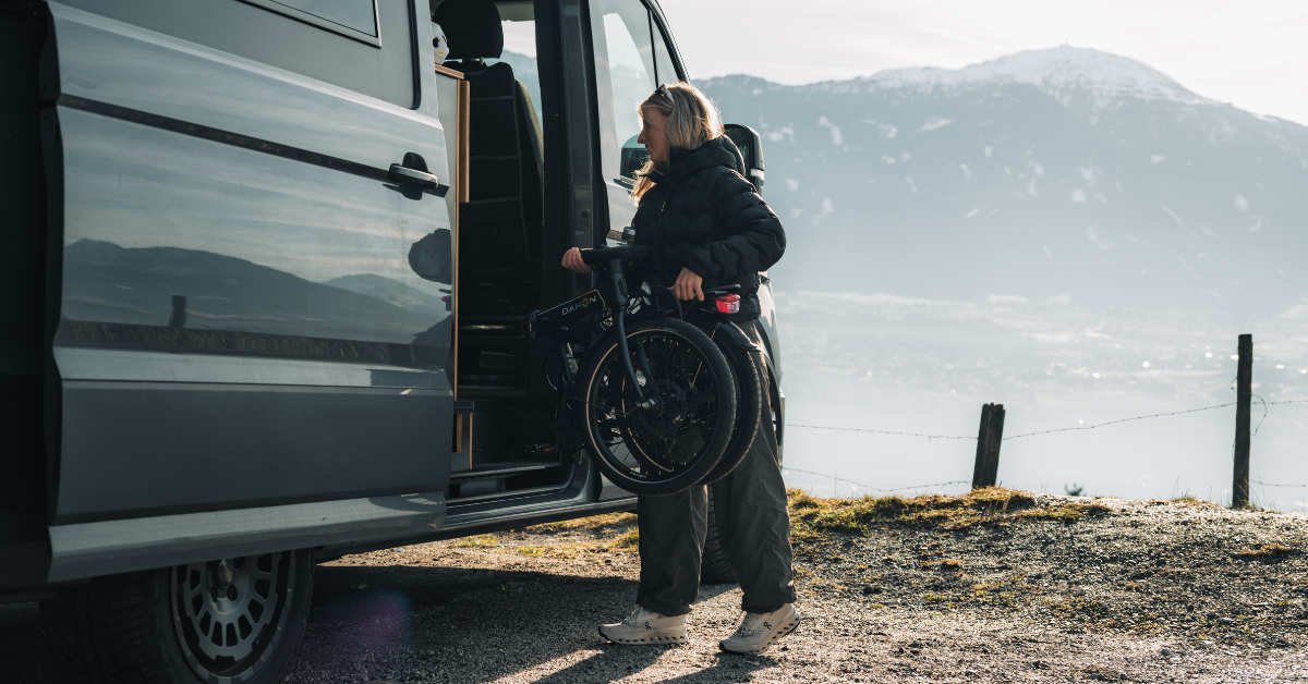 A person loads a folded black bicycle into the side door of a van, with mountains and water in the background.
