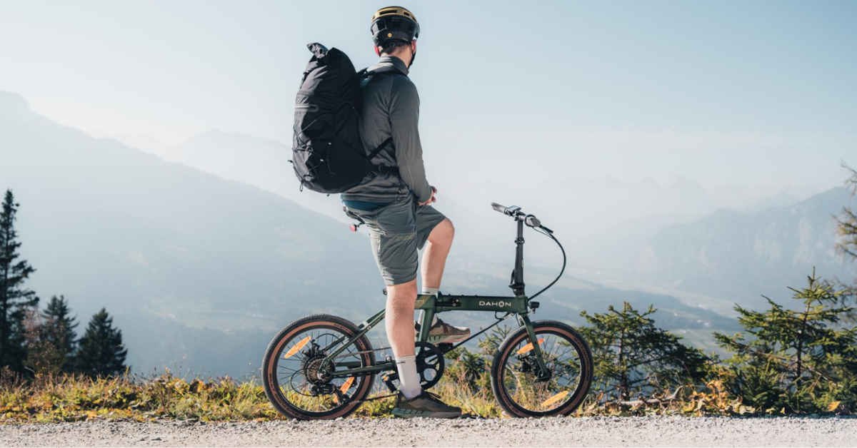 A cyclist stands with a folding bike, looking out over a mountain landscape.