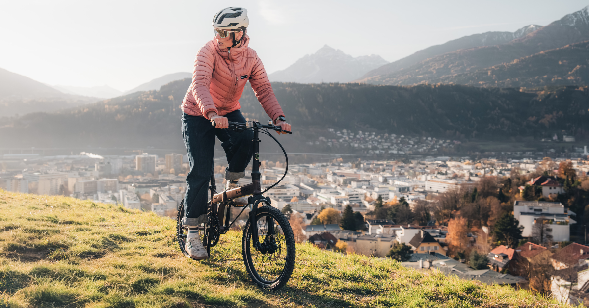 A person in a pink jacket rides a folding bike on a grassy hill, with a valley city in the background.