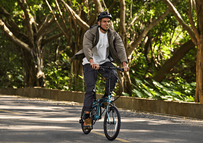 Teal DAHON Mu Ex folding bike in motion with a rider on a tree-lined road.